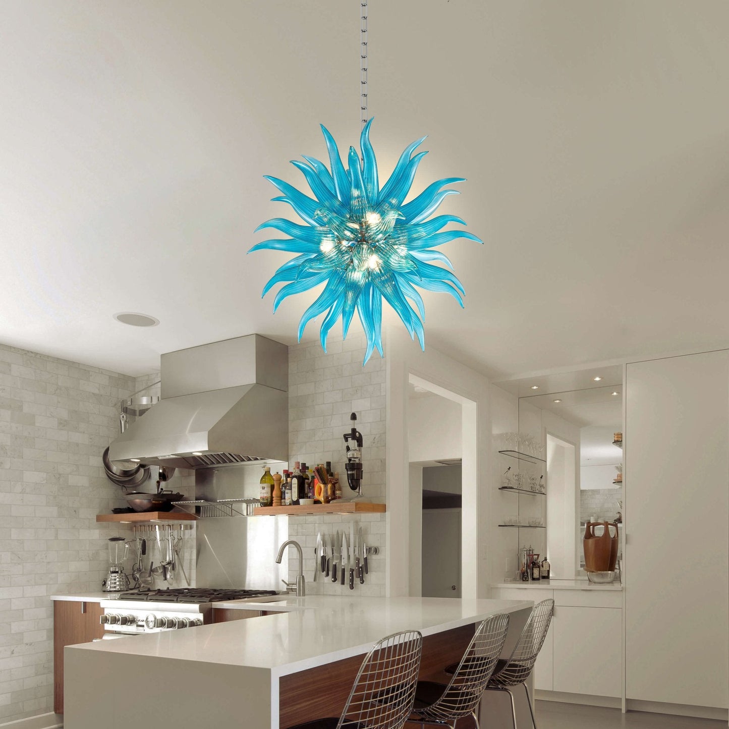 Modern ice-blue glass art chandelier centered over a contemporary kitchen island with white cabinetry and minimalist bar stools.
