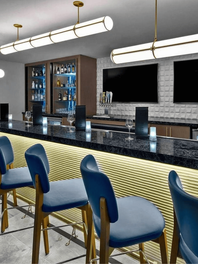 Luxury home bar or hotel lounge featuring long linear tube pendant lighting above blue velvet bar stools.
