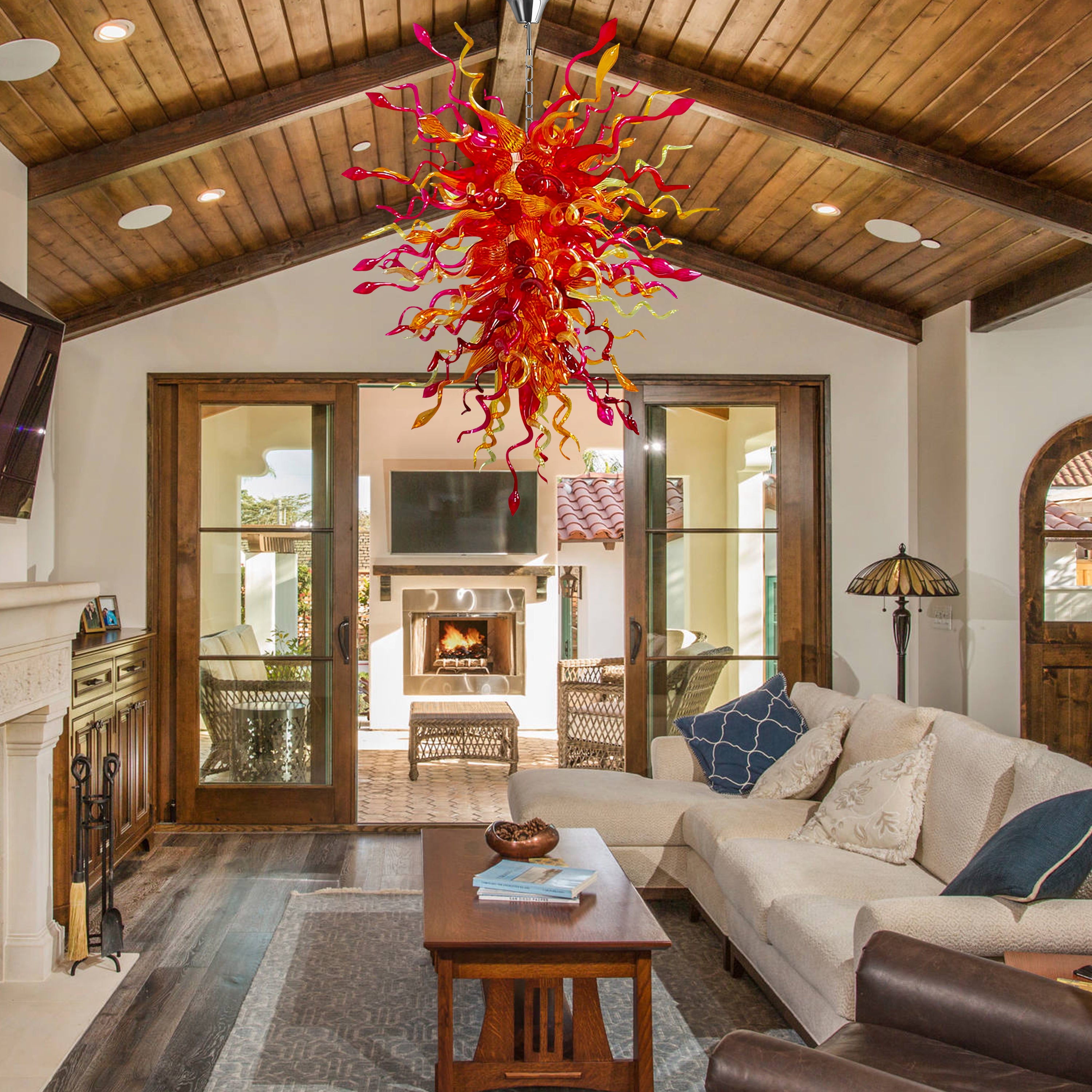 Hand blown glass chandelier with red and yellow hues hanging from a vaulted wooden ceiling in a cozy living room with a fireplace.