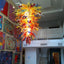 Colorful blown glass chandelier with an explosion of swirling glass in yellow, red, orange, and blue, displayed in an interior setting with artwork on the walls.
