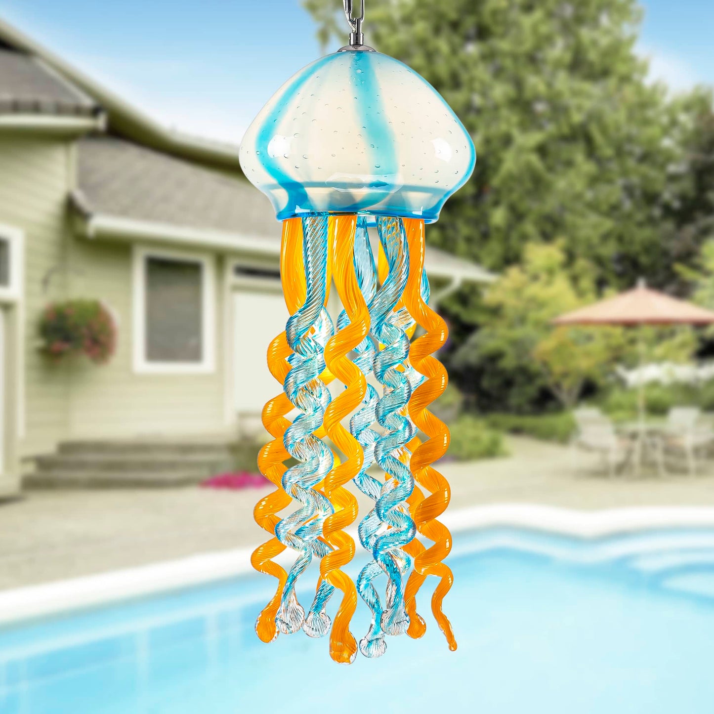 Blue and orange glass jellyfish wind chime hanging by a swimming pool in a bright sunny garden.