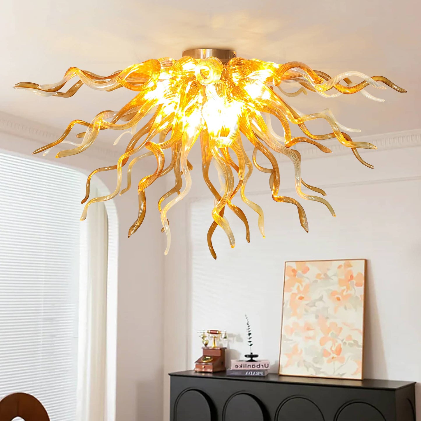 Close-up shot of an illuminated radiant hand-blown glass ceiling chandelier featuring twisted amber and light champagne glass curls, casting a golden glow over a modern black hallway console table with an abstract art piece.