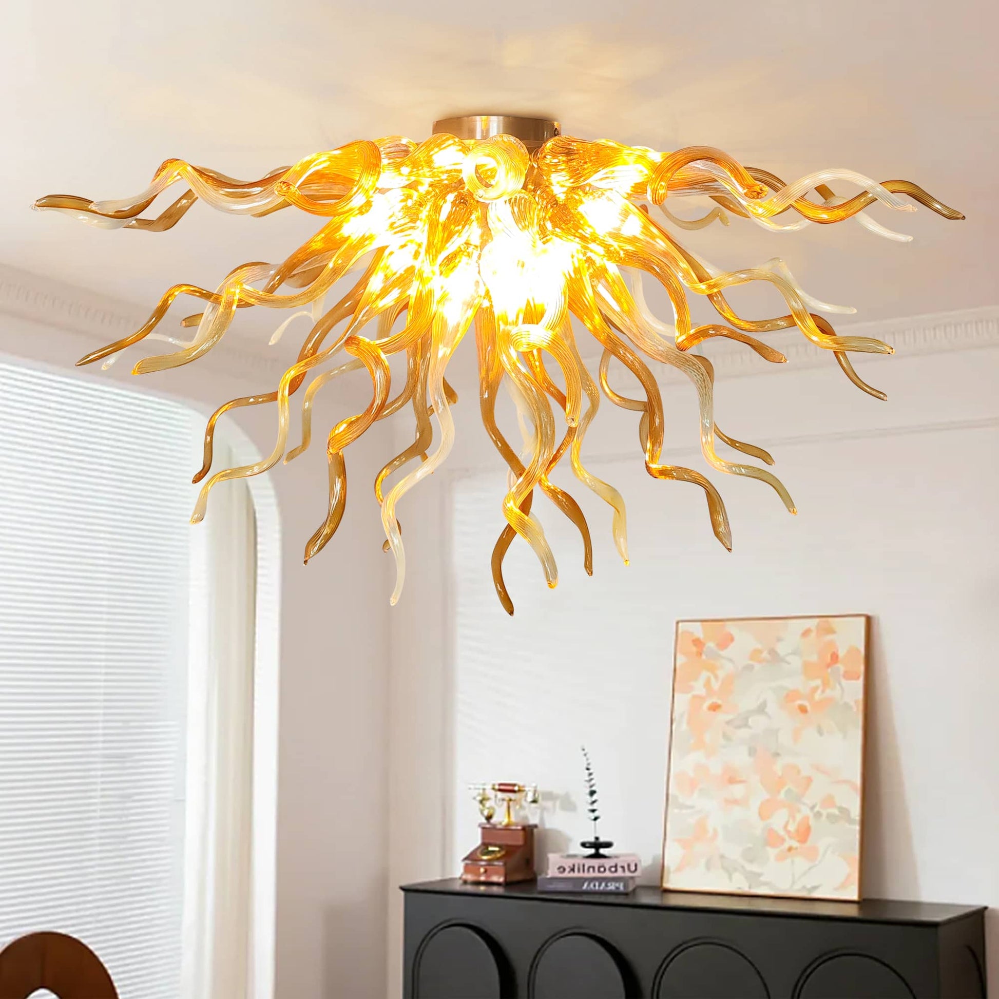 Close-up shot of an illuminated radiant hand-blown glass ceiling chandelier featuring twisted amber and light champagne glass curls, casting a golden glow over a modern black hallway console table with an abstract art piece.