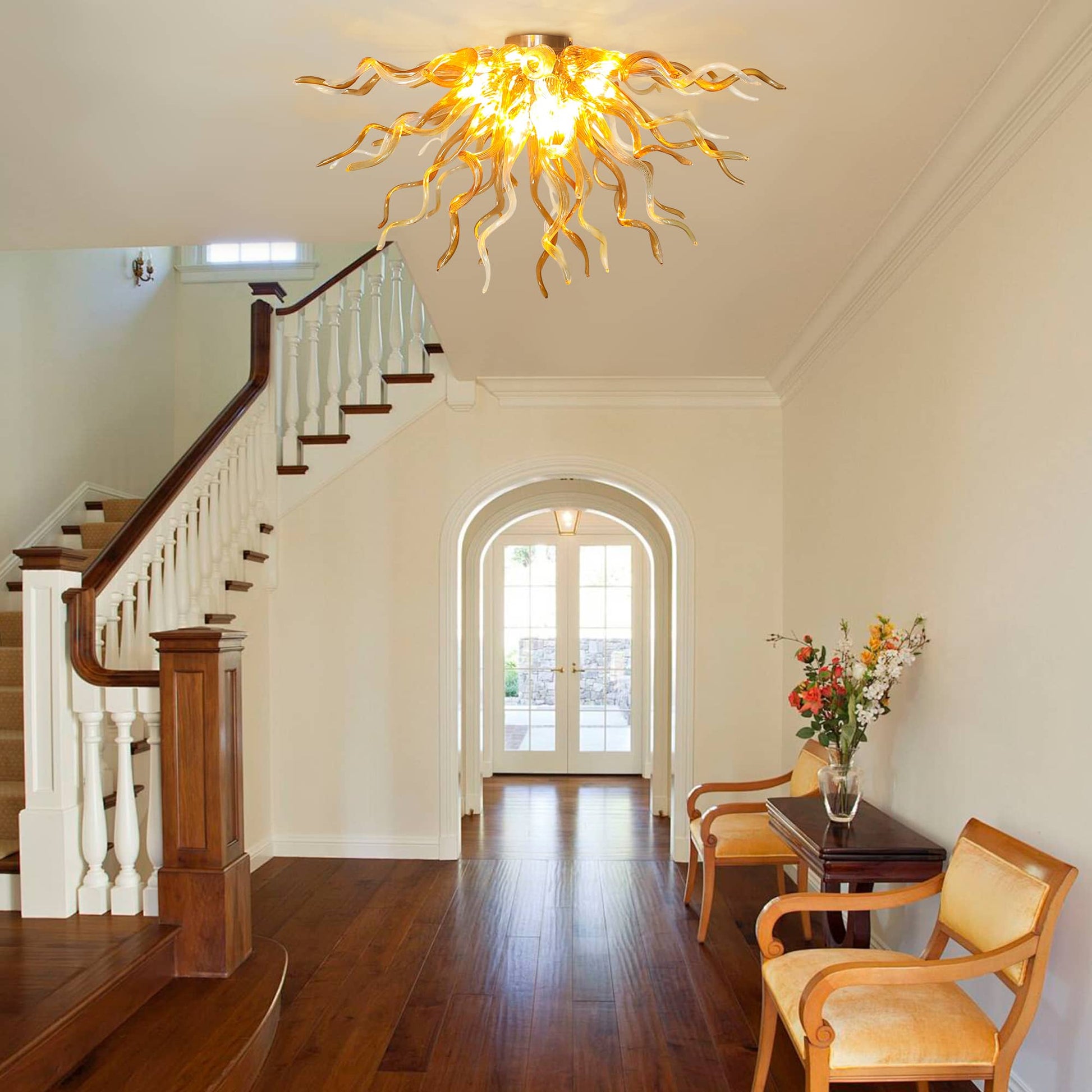 Grand home entryway foyer scene with a warm golden glass branching chandelier casting light across a polished hardwood floor next to a white wooden staircase.