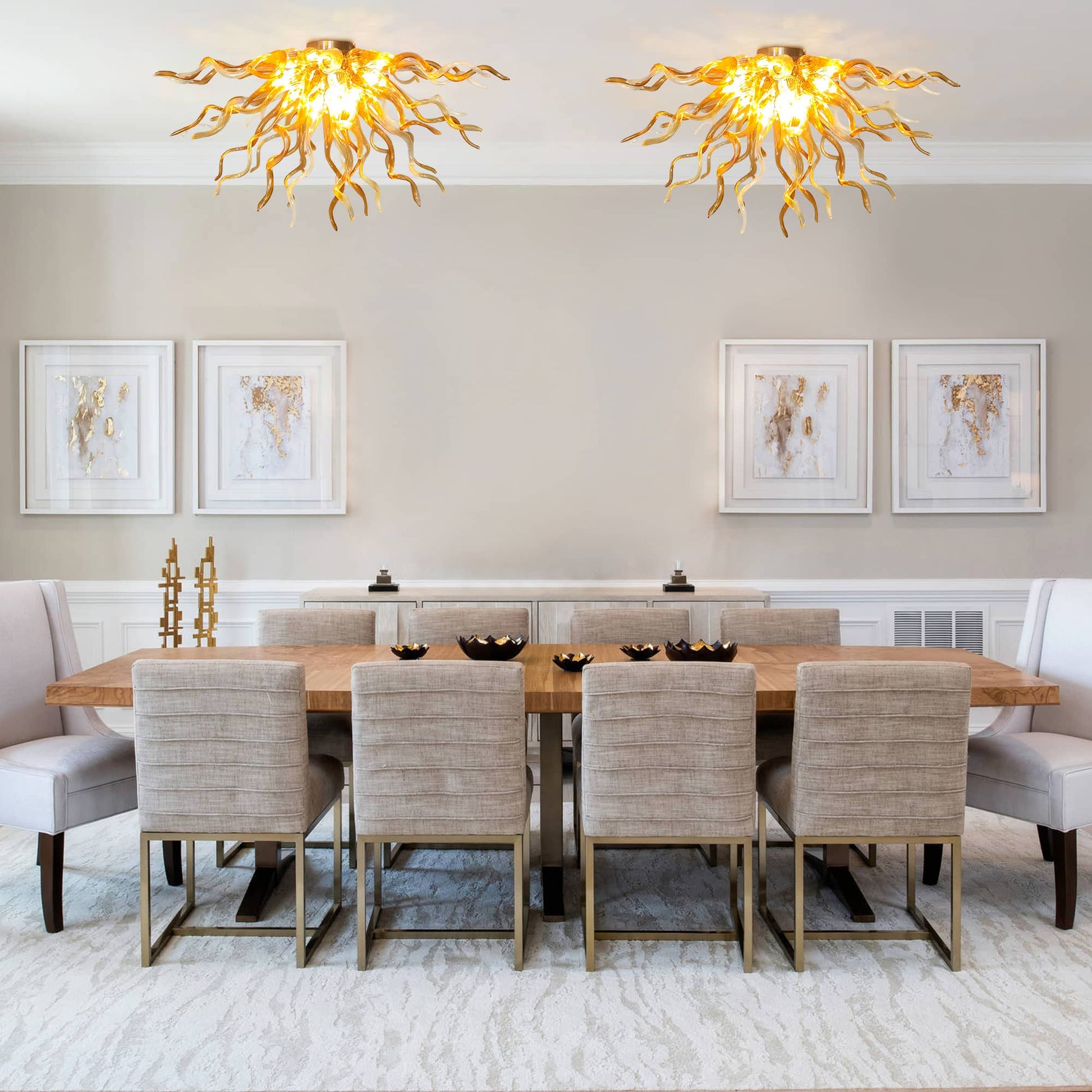 Upscale formal dining room setting featuring two identical hand-blown amber glass chandeliers installed side-by-side over a long dining table with cream tufted chairs.