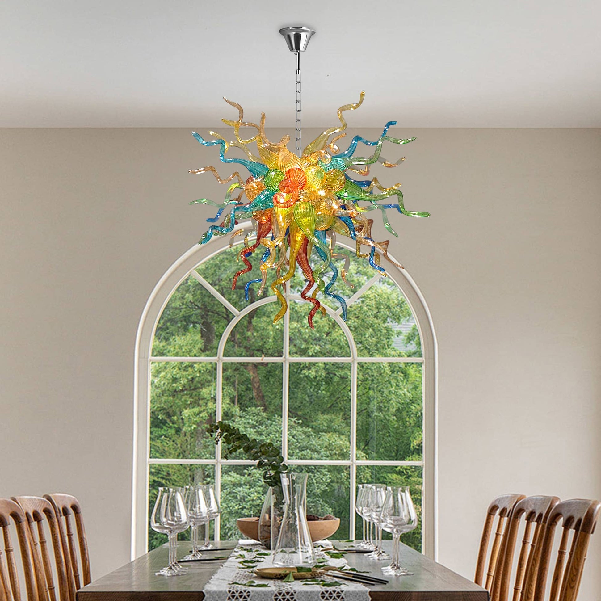 Artistic glass chandelier hanging over a wooden dining table with a large arched window in the background.