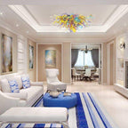 Modern luxury living room with white sofas and blue accents, showcasing the long colorful glass sculptural chandelier as a central ceiling focal point.