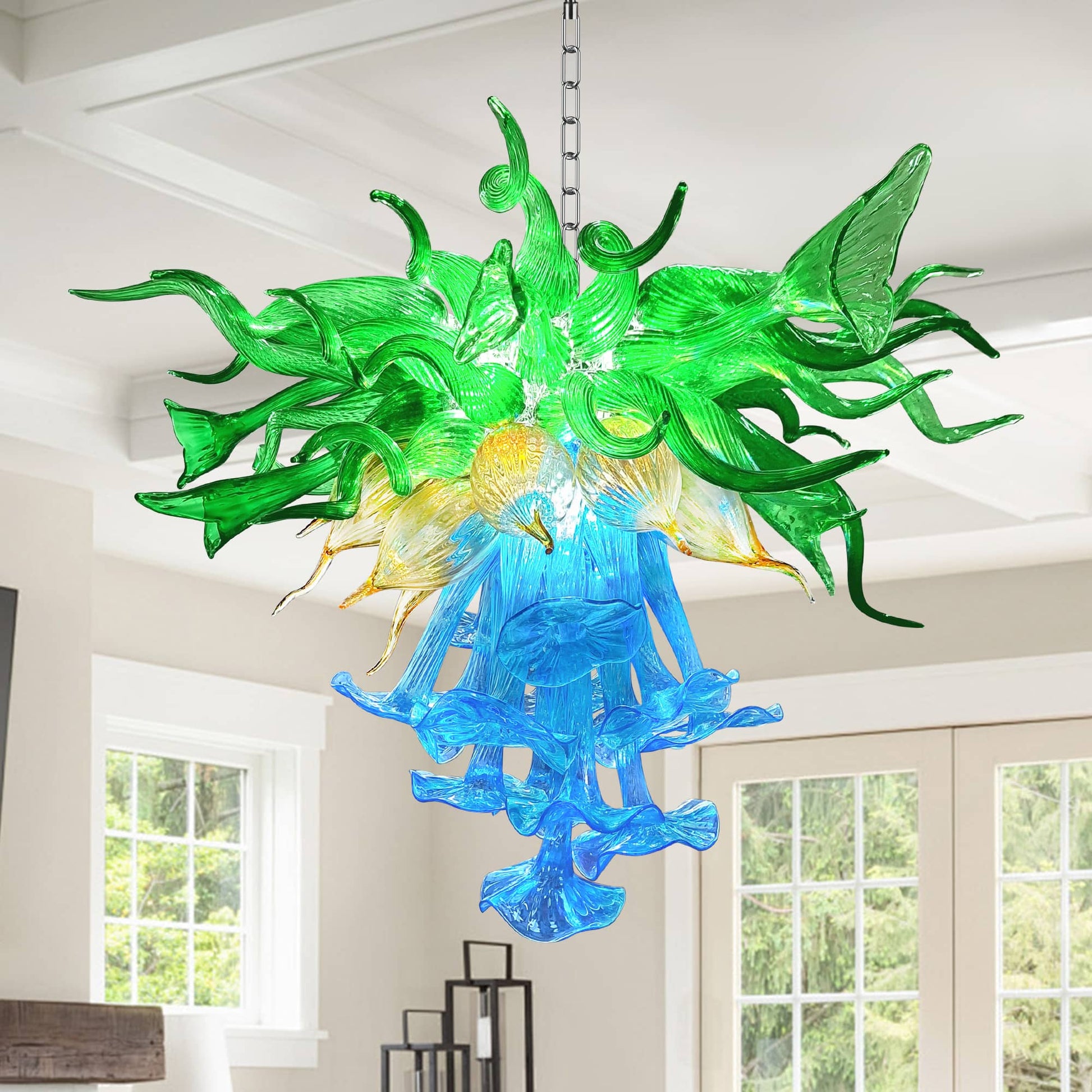 Green and blue hand-blown glass chandelier hanging in a modern living room with white walls and large windows.