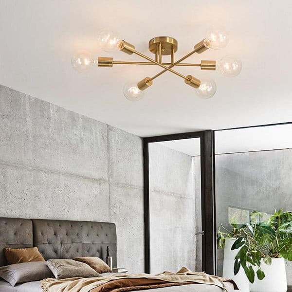Sputnik linear brass ceiling chandelier illuminating a modern minimalist bedroom with a grey tufted headboard.