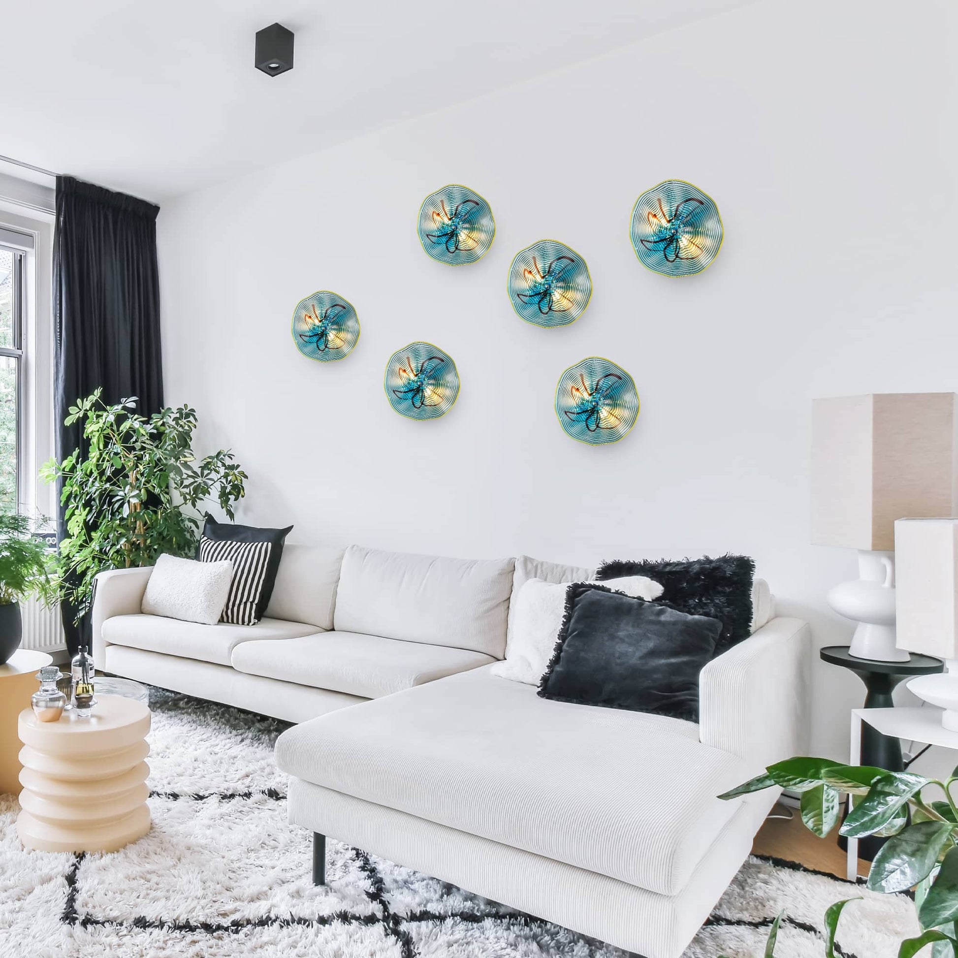 Six hand-blown aqua glass wall plates arranged in a floral cluster above a white luxury sofa.